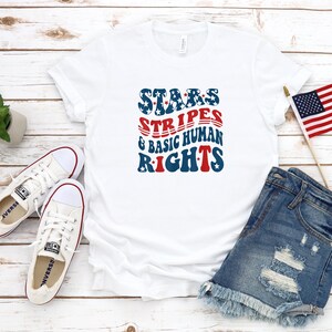 May include: White t-shirt with the text "STARS, STRIPES & BASIC HUMAN RIGHTS" in red, white, and blue. The image also includes white sneakers, denim shorts, a small American flag, and a small potted plant.