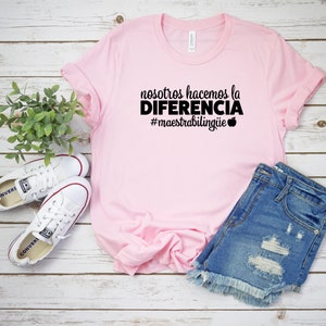 Puede incluir: Una camiseta rosa claro con el texto en espa&ntilde;ol "nosotros hacemos la DIFERENCIA #maestrabiling&uuml;e" en negro. La camiseta se combina con pantalones cortos de mezclilla y zapatillas blancas. Una ramita de hojas verdes est&aacute; en el fondo.