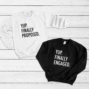 May include: Two sweatshirts, one white and one black, with the phrases "YUP. FINALLY PROPOSED." and "YUP. FINALLY ENGAGED." printed in black and white, respectively. The sweatshirts are displayed on a white wooden surface.