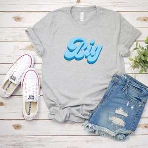 May include: A gray t-shirt with the word "Big" in a blue retro font. The shirt is laid out on a white wooden surface with a pair of white Converse sneakers and a pair of blue denim shorts.