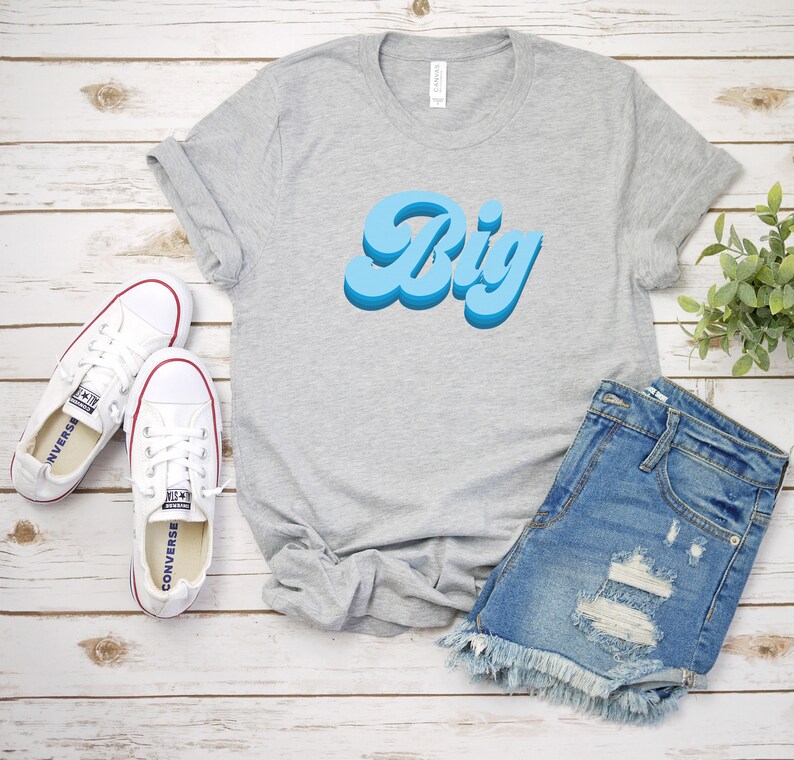 May include: A gray t-shirt with the word "Big" in a blue retro font. The shirt is laid out on a white wooden surface with a pair of white Converse sneakers and a pair of blue denim shorts.