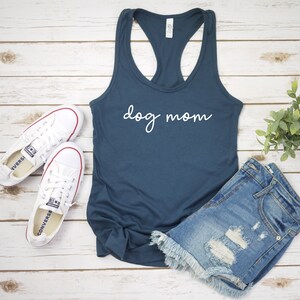 May include: A navy blue tank top with the text "dog mom" in white lettering. The tank top is laying on a white wooden surface with a pair of white sneakers and a pair of denim shorts.