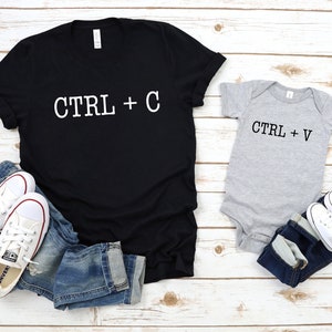 May include: A black t-shirt with the text "CTRL + C" in white lettering and a grey baby onesie with the text "CTRL + V" in black lettering. Both shirts are on a white wooden surface with blue jeans and white sneakers.