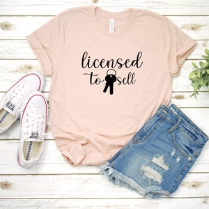 Puede incluir: Una camiseta rosa con el texto "licensed to sell" y un gr&aacute;fico de llave negra. La camiseta est&aacute; sobre una superficie de madera blanca con un par de zapatillas blancas y pantalones cortos de mezclilla azul.