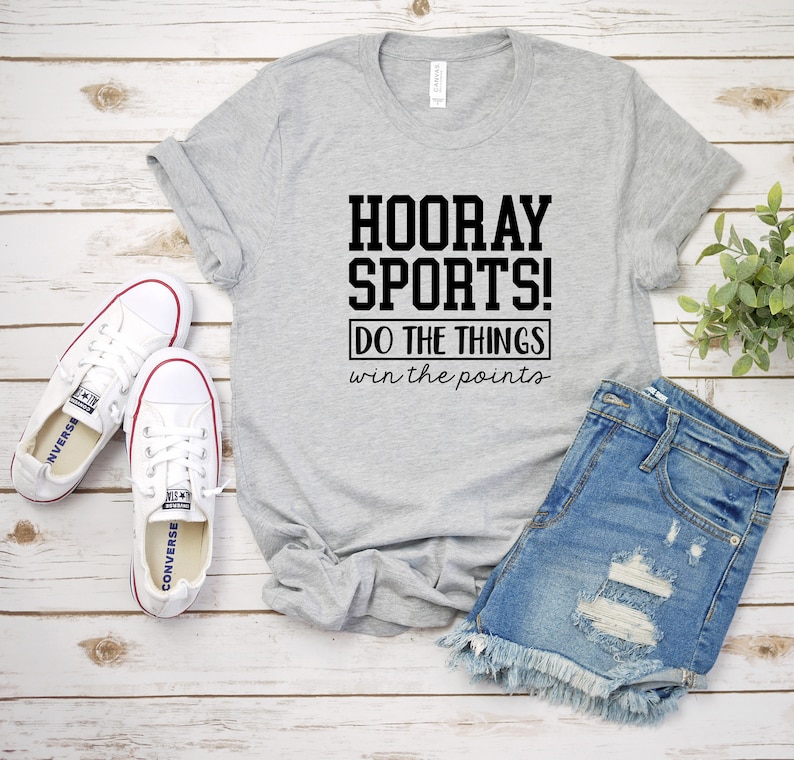 Op de afbeelding: Een grijs T-shirt met de tekst "HOORAY SPORTS! DO THE THINGS win the points" in zwart gedrukt. Het shirt ligt op een houten oppervlak met een paar witte sneakers en een blauwe spijkerbroek.