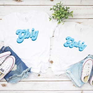 May include: Two white t-shirts with the word "Y'all Big" written in blue, 3D-style lettering. The shirts are laid out on a white wooden surface with denim shorts and white sneakers.