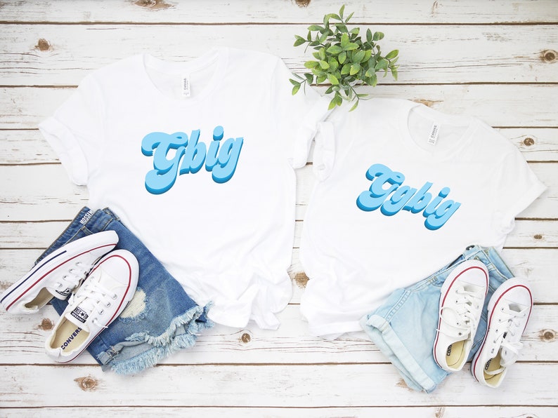 May include: Two white t-shirts with the word "Y'all Big" written in blue, 3D-style lettering. The shirts are laid out on a white wooden surface with denim shorts and white sneakers.