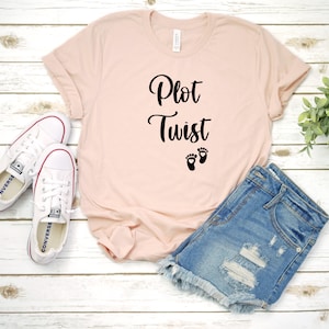 May include: A pink t-shirt with the text "Plot Twist" and a pair of black baby footprints. The shirt is laying on a white wooden surface with a pair of white sneakers and a pair of blue denim shorts.