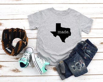 May include: A gray t-shirt with a black outline of the state of Texas and the text "made." printed on it. The shirt is laid out on a white wooden surface with a baseball glove, baseball, and a pair of blue jeans.