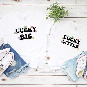 Op de afbeelding: Twee witte T-shirts met de tekst "Lucky Big" en "Lucky Little" in zwart met kleurrijke klavertjevier-accenten. De shirts liggen op een wit houten oppervlak met denim shorts en witte sneakers.