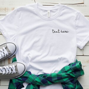 May include: White t-shirt with the text "text here" embroidered on the front. The shirt is laid flat on a white wooden surface with a green and blue plaid shirt and gray sneakers.