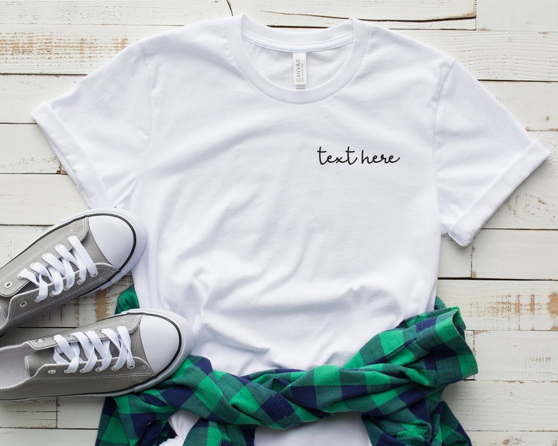 May include: White t-shirt with the text "text here" embroidered on the front. The shirt is laid flat on a white wooden surface with a green and blue plaid shirt and gray sneakers.