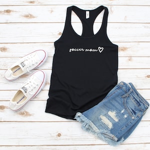 May include: Black tank top with the text "soccer mom" and a white heart on the front. The tank top is paired with denim shorts and white sneakers.