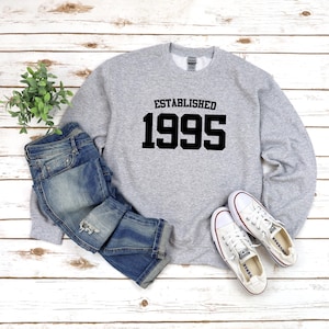 May include: Gray sweatshirt with black text that reads "Established 1995". The sweatshirt is paired with blue jeans and white sneakers.