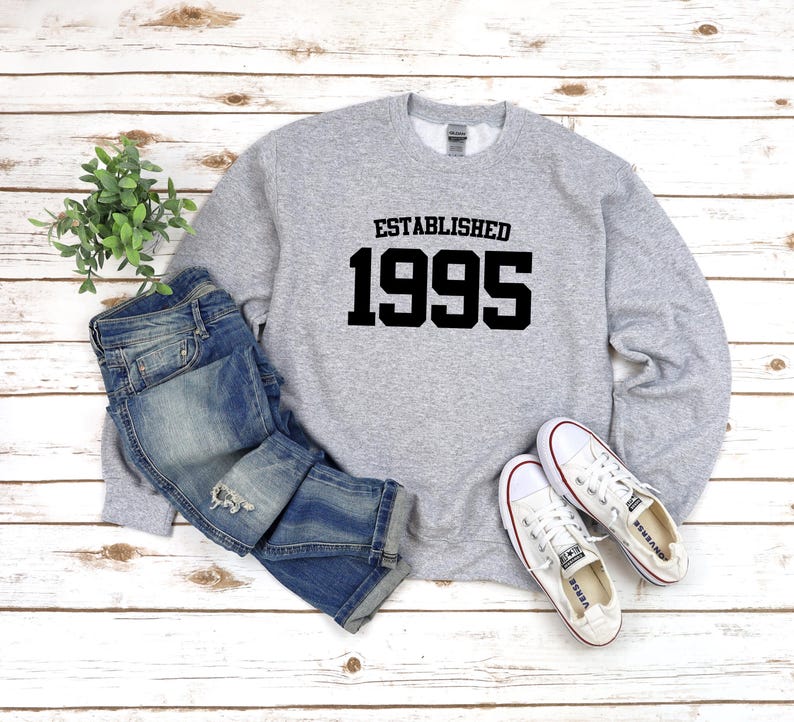 May include: Gray sweatshirt with black text that reads "Established 1995". The sweatshirt is paired with blue jeans and white sneakers.