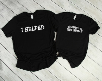 May include: Two black t-shirts with white text. One shirt says "I Helped" and the other says "Growing a Tiny Human".