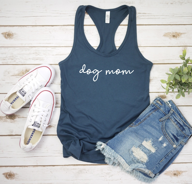 May include: A navy blue tank top with the text "dog mom" in white script lettering. The tank top is laying on a white wooden surface with a pair of white sneakers and a pair of blue denim shorts.