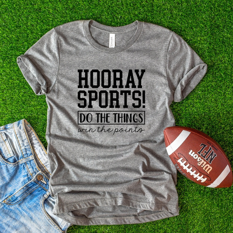 Op de afbeelding: Een grijs T-shirt met de tekst "HOORAY SPORTS! DO THE THINGS win the points" erop gedrukt. Het shirt ligt op een groene ondergrond met een bruine voetbal ernaast.