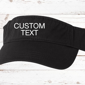 May include: Black visor with the words "CUSTOM TEXT" in white block letters. The visor is made of a solid black material and has a curved brim. The background is a white wooden surface.