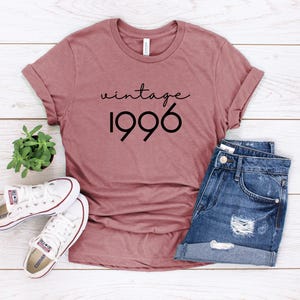 May include: A mauve short-sleeved t-shirt with the text vintage 1996 printed in black, paired with blue denim shorts and white canvas sneakers. The shirt features a crew neckline and rolled sleeves, creating a casual look with these items.