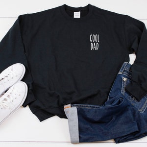 Op de afbeelding: Zwart sweatshirt met de tekst "COOL DAD" in wit op de voorkant. Het sweatshirt ligt op een wit oppervlak met een paar witte sneakers en een blauwe jeans.