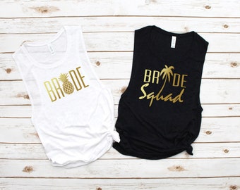 May include: Two sleeveless tank tops, one white and one black, with gold text. The white tank top says "BRIDE" with a pineapple graphic. The black tank top says "BRIDE Squad" with a palm tree graphic. Both tops are knotted at the bottom.
