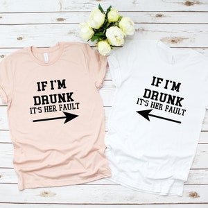 Op de afbeelding: Twee T-shirts met een bijpassend ontwerp. Het roze T-shirt zegt "Als ik dronken ben, is het haar schuld" met een pijl die naar rechts wijst. Het witte T-shirt zegt "Als ik dronken ben, is het haar schuld" met een pijl die naar links wijst.