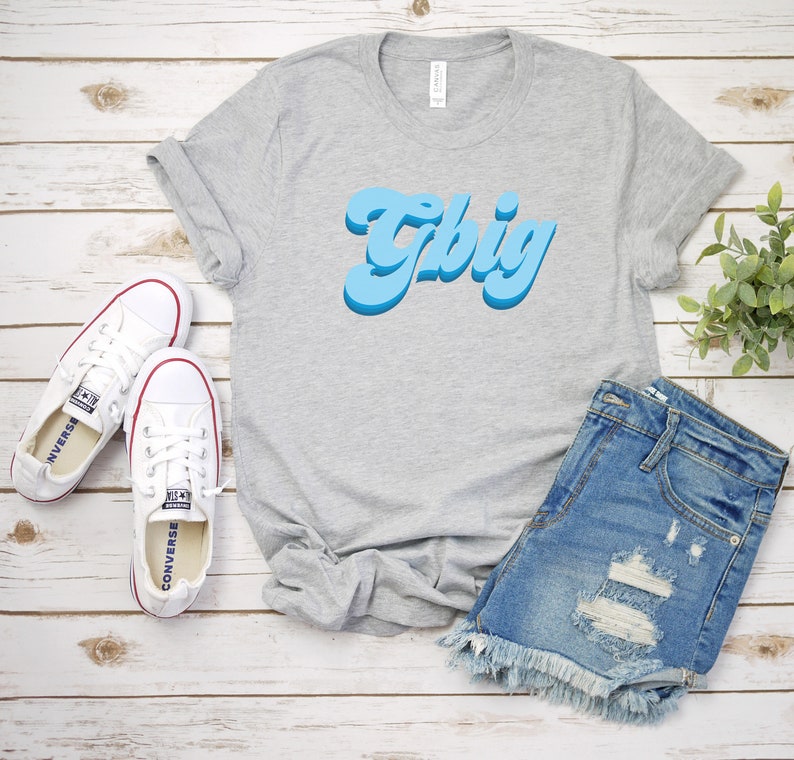 May include: A gray t-shirt with a blue retro-style graphic that says "Tbig". The shirt is paired with denim shorts and white sneakers.