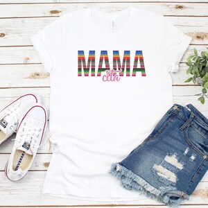 Op de afbeelding: Wit T-shirt met de tekst "MAMA cita" in kleurrijke gestreepte letters. De tekst is in een kleurrijk gestreept patroon dat doet denken aan een Mexicaanse deken. Het shirt ligt plat op een wit houten oppervlak met een paar witte sneakers, een spijkerbroekje en een potplant.