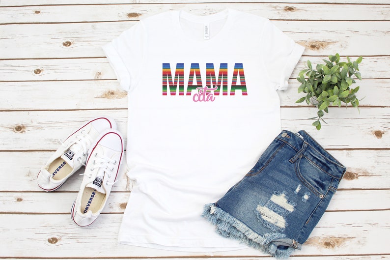 Op de afbeelding: Wit T-shirt met de tekst "MAMA cita" in kleurrijke gestreepte letters. De tekst is in een kleurrijk gestreept patroon dat doet denken aan een Mexicaanse deken. Het shirt ligt plat op een wit houten oppervlak met een paar witte sneakers, een spijkerbroekje en een potplant.