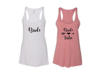 May include: Two tank tops for a bridal party. One white tank top says "Bride" in black script. The other pink tank top says "Bride Tribe" in black script with an arrow and heart.