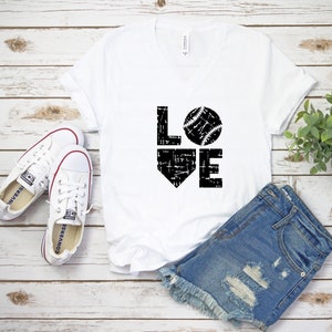 May include: White V-neck t-shirt with the word "LOVE" in black, featuring a baseball graphic. The shirt is paired with denim shorts and white canvas sneakers. A sprig of greenery is in the background.