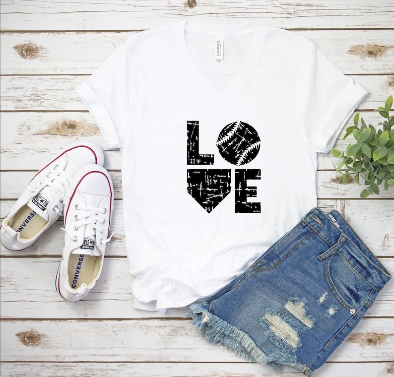 May include: White V-neck t-shirt with the word "LOVE" in black, featuring a baseball graphic. The shirt is paired with denim shorts and white canvas sneakers. A sprig of greenery is in the background.
