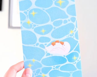 May include: A postcard featuring a whimsical illustration of a white swan with orange accents, floating on a light blue water-like background with yellow star accents. The postcard is held by a hand.