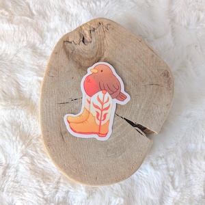 Op de afbeelding: Een sticker met een cartoon roodborstje dat een cowboylaars draagt met een bloemenpatroon. Het roodborstje is bruin met een rode borst en zit bovenop de laars.