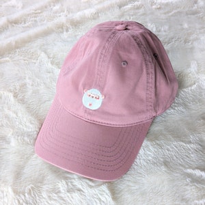 Può includere: Un berretto da baseball rosa con un designo di pecora ricamato in bianco. Il berretto ha una visiera curva e una forma tradizionale di berretto da baseball.
