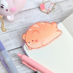 May include: A clear acrylic charm featuring a cartoon illustration of a light orange bear sleeping on its back. The bear has a small, pink nose and closed eyes.