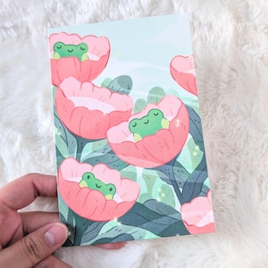 Op de afbeelding: Een ansichtkaart met een illustratie van groene kikkers in roze bloemen. De achtergrond heeft een lichtblauwe lucht en groen gebladerte. Het ontwerp is grillig en kleurrijk.