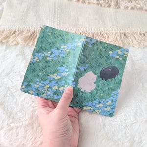 May include: A notebook cover featuring a whimsical illustration of two cats in a field of grass and blue flowers. The cover is held open, revealing the interior. The design includes the text "arcdesign" in the lower left corner.