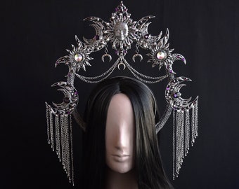 May include: Silver-toned celestial crown featuring sun and moon motifs, adorned with purple accents and dangling chains. The crown is displayed on a mannequin head with long, black hair against a black backdrop. A unique headpiece.