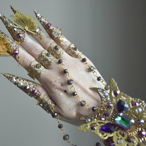 May include: A set of gold-toned finger rings with intricate designs and butterfly accents. The rings feature iridescent gemstones and are connected by a chain with beads.