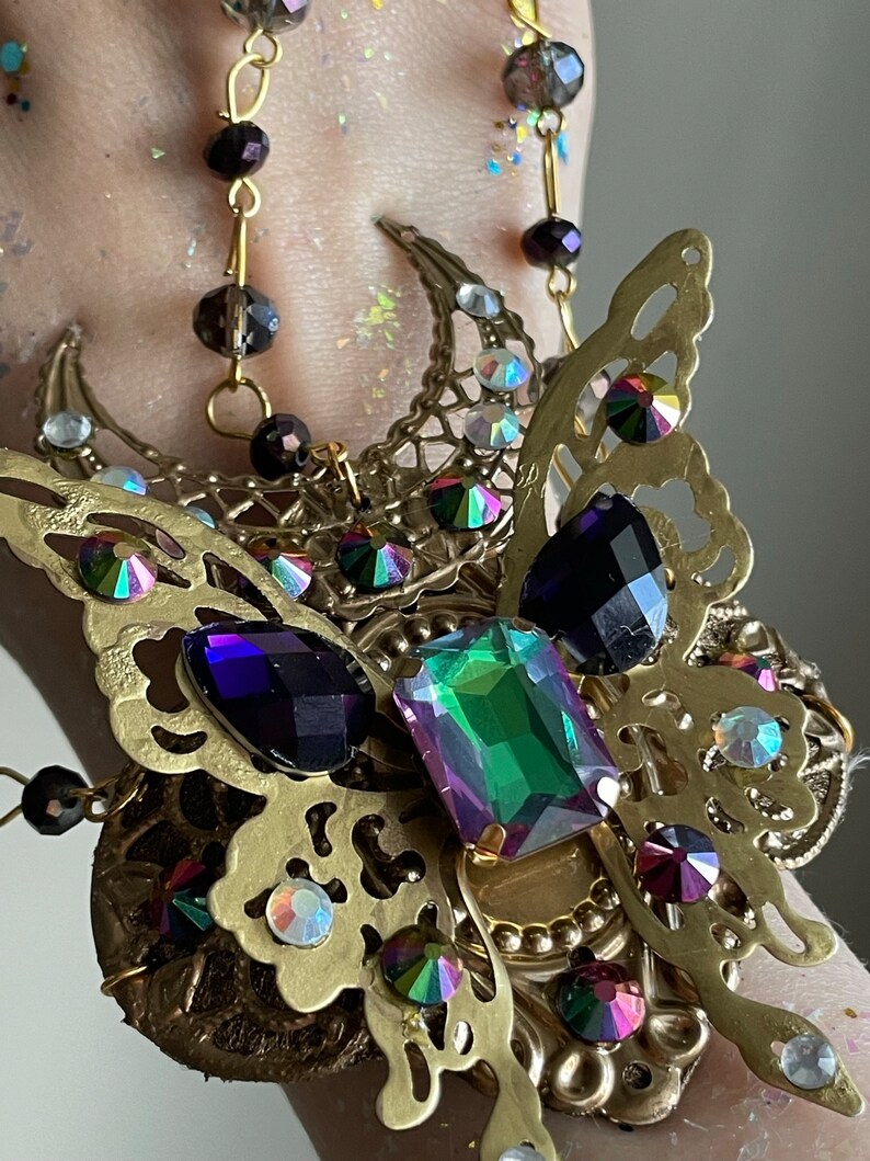 May include: A gold-toned butterfly-shaped bracelet with iridescent and purple gemstones. The bracelet features a delicate, intricate design and is adorned with a variety of crystals.