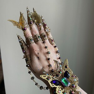 May include: A gold-toned finger ring set with a butterfly design. The set includes multiple rings with intricate details and a chain connecting them. The rings are adorned with iridescent gemstones and crystals.