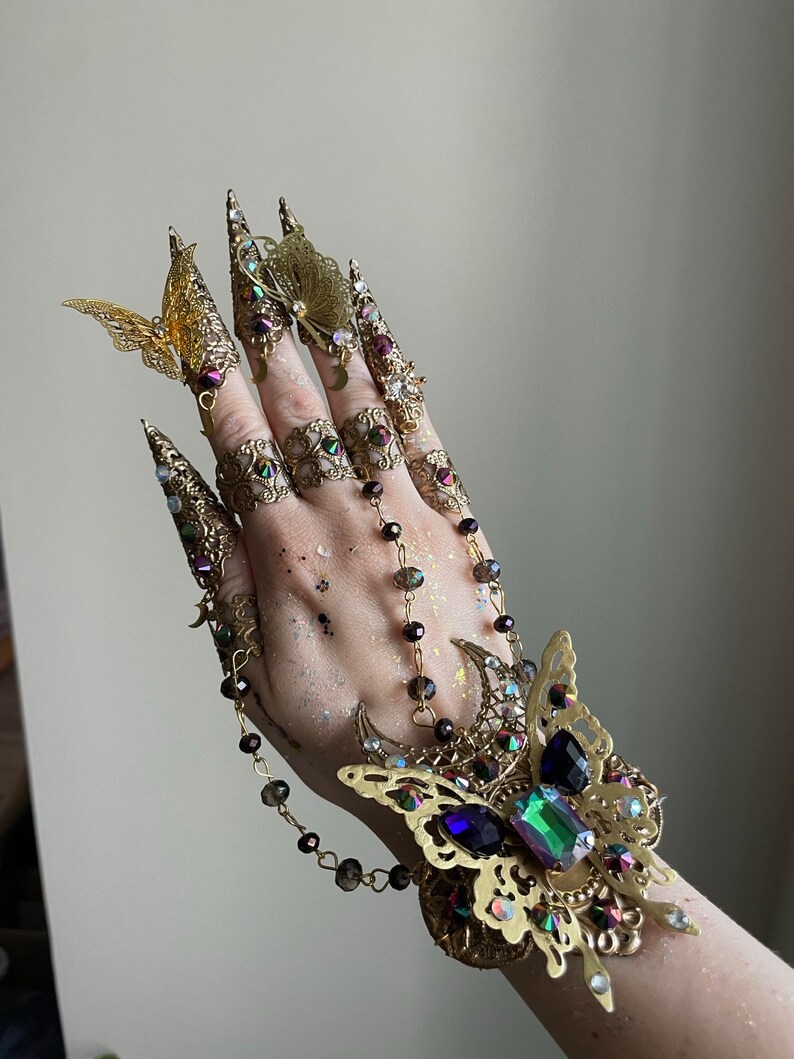 May include: A gold-toned finger ring set with a butterfly design. The set includes multiple rings with intricate details and a chain connecting them. The rings are adorned with iridescent gemstones and crystals.