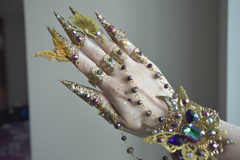 May include: A set of gold-toned finger rings with intricate designs and butterfly accents. The rings feature iridescent gemstones and are connected by a chain with beads.