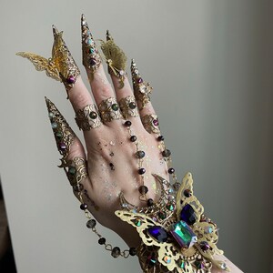 May include: A gold-toned, ornate hand jewelry set with butterfly and crescent moon motifs. The set includes finger rings, a bracelet, and a chain connecting the pieces. The jewelry is adorned with iridescent and clear crystals.