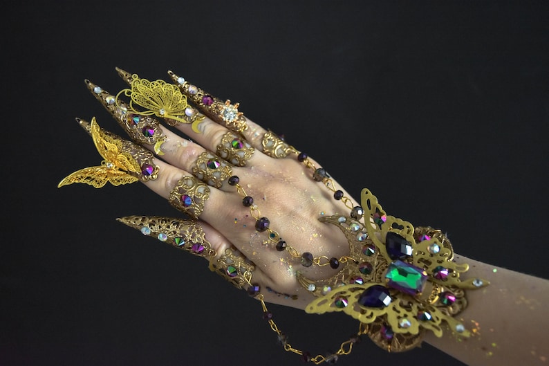 May include: A gold-toned hand jewelry set with intricate butterfly designs and colorful gemstones. The set includes rings, a bracelet, and long, pointed finger coverings.