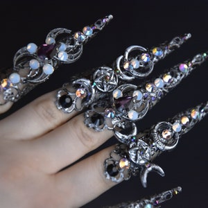 May include: Five silver rings with pointed tips, adorned with iridescent crystals, black stones, and white opalescent stones. The rings feature crescent moon and pentagram designs.