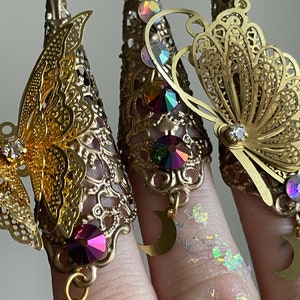May include: Gold-toned finger rings with intricate butterfly designs and dangling crescent moon charms. The rings are adorned with colorful rhinestones.