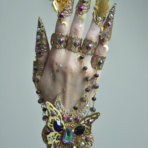 May include: A gold-toned finger ring set with a butterfly design and iridescent gemstones. The set includes multiple rings with a crescent moon design and a chain with iridescent beads.